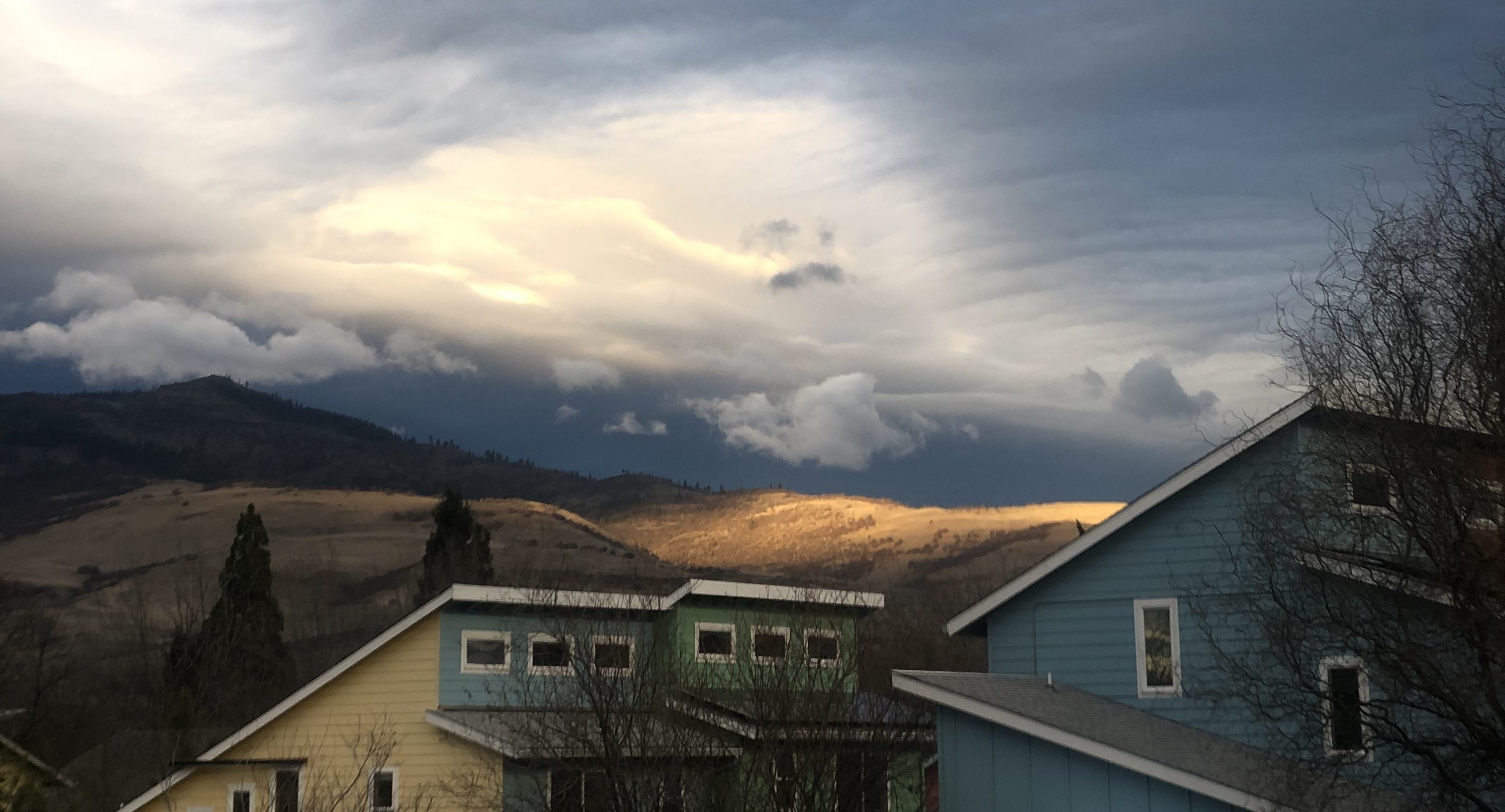 A hillside under a dramatic cloudscape is lit by one patch of sunlight. A few houses are dark in the foreground.